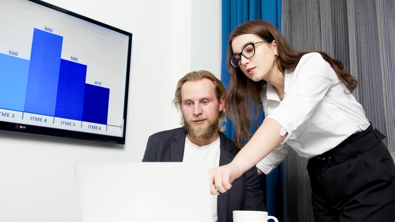 A Woman Standing Beside a Man Sitting in Front of a White Laptop